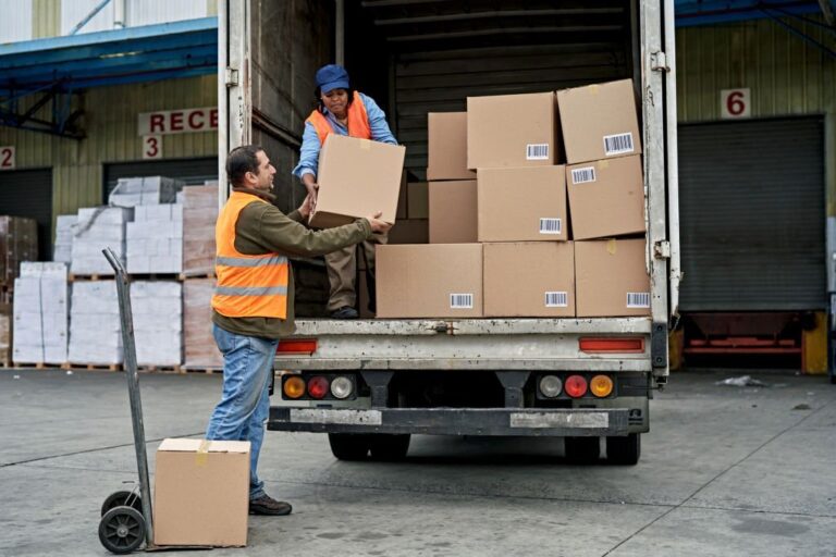 Full length view of late 40s man moving boxes from loading dock with hand truck and lifting into hands of woman organizing merchandise on vehicle.